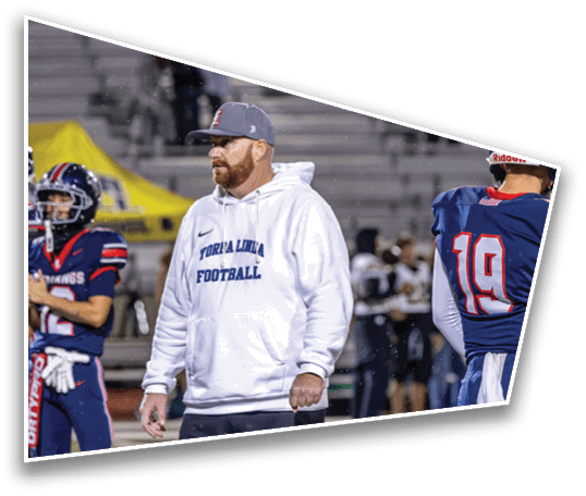 People in football uniforms and a person in a white hoodie standing on a field during a game or practice. Description generated by AI