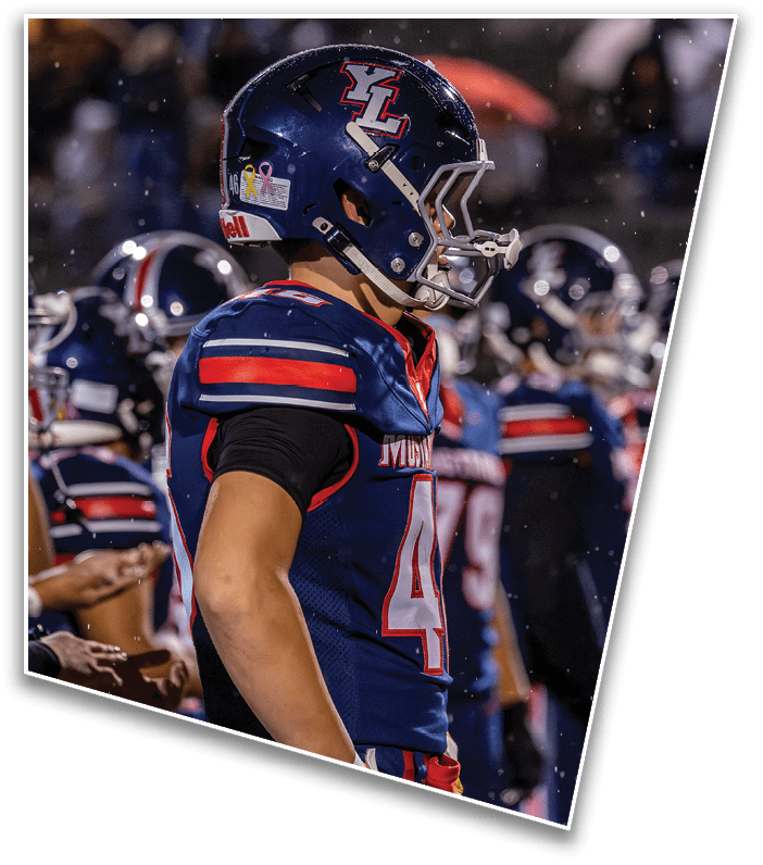 A person wearing a blue football uniform and helmet stands on the sidelines with other players in the background during a nighttime game. Description generated by AI