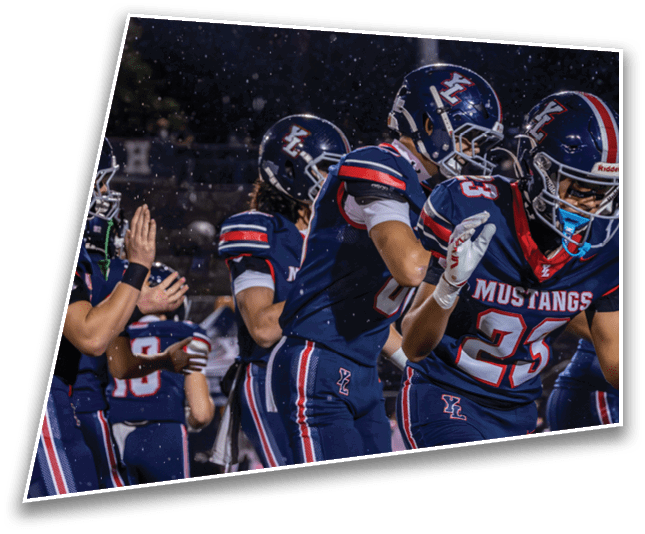 People wearing football helmets and uniforms stand together on a field at night under bright stadium lights with visible raindrops falling. Description generated by AI
