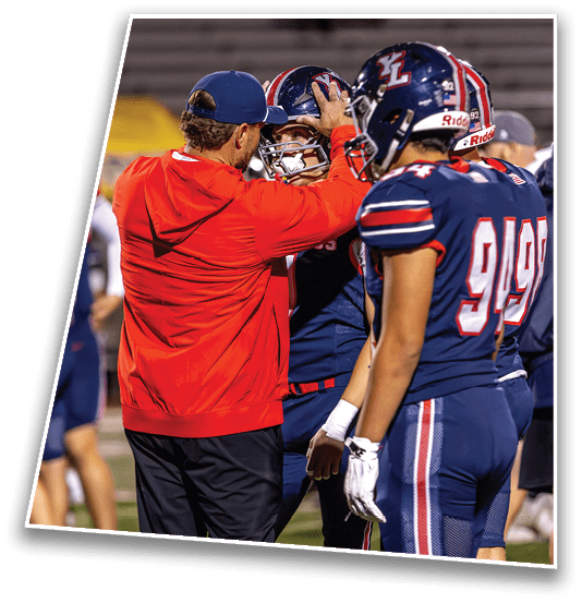 A person in a red jacket and blue cap is adjusting the helmet of a person in a blue football uniform while another person in a similar uniform stands nearby on a sports field. Description generated by AI