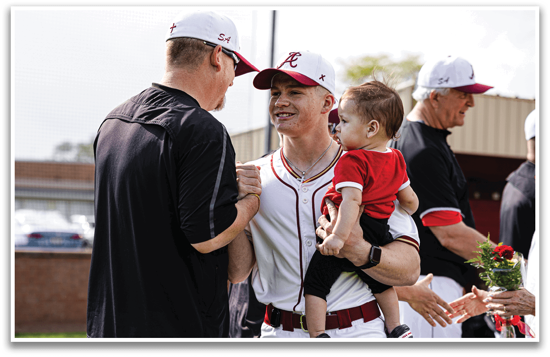 A man wearing a red and white baseball uniform is holding a baby. Description generated by AI