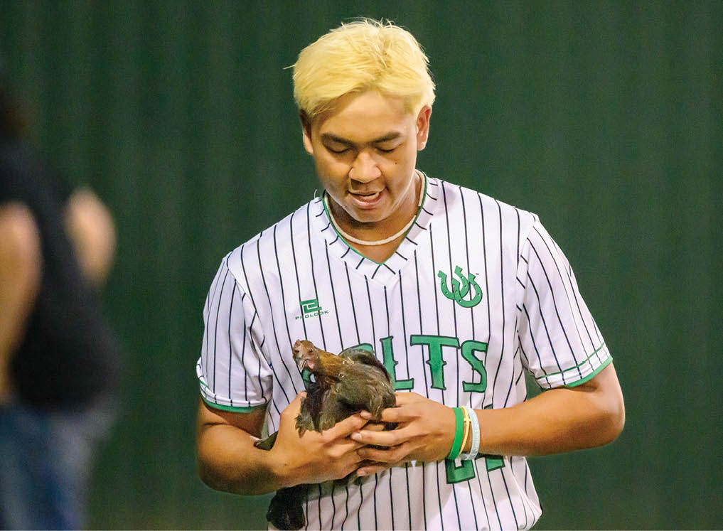A man wearing a baseball uniform is holding a chicken. Description generated by AI