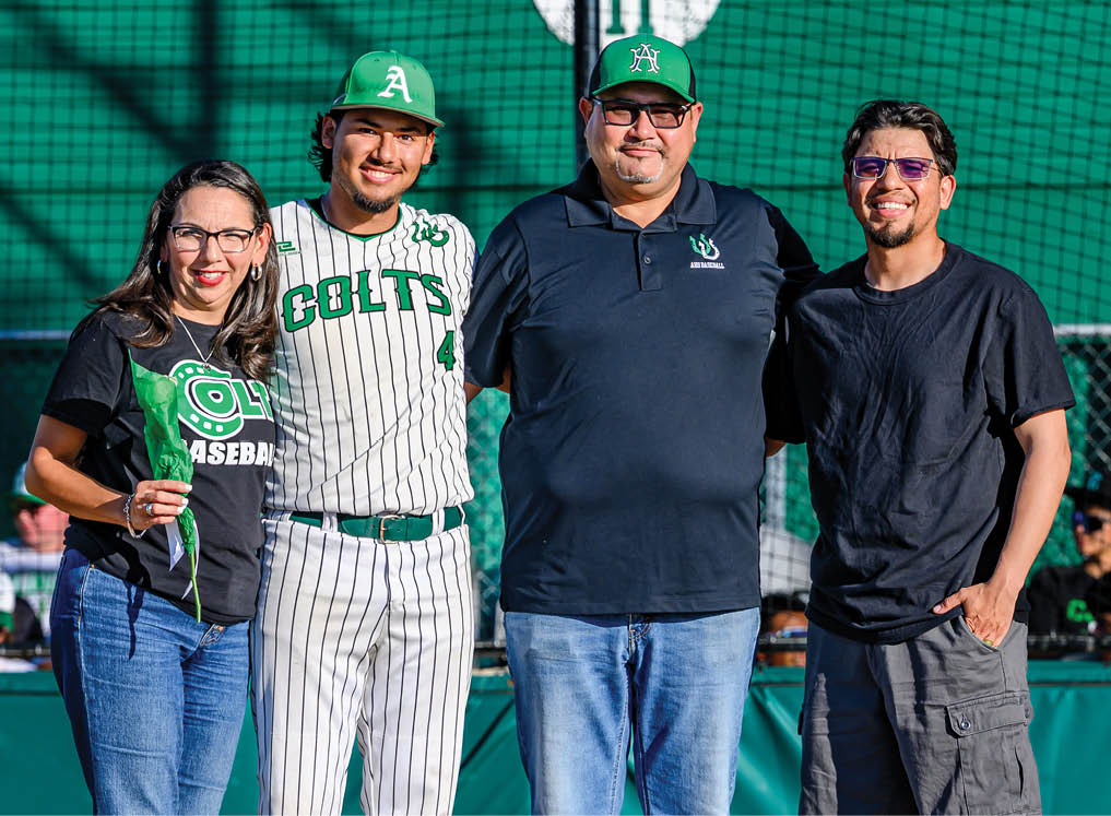 A man wearing a baseball uniform stands between two men and a woman. Description generated by AI
