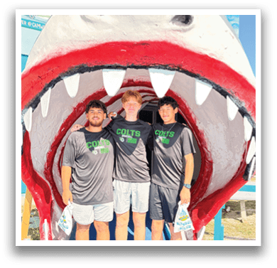 Three men wearing grey shirts with the word “Colts" on them pose for a picture in front of a large shark sculpture. AI generated content