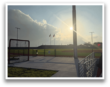 A person is standing on a sidewalk near a baseball field. The sun is shining brightly, and there is a flag on a pole. AI generated content