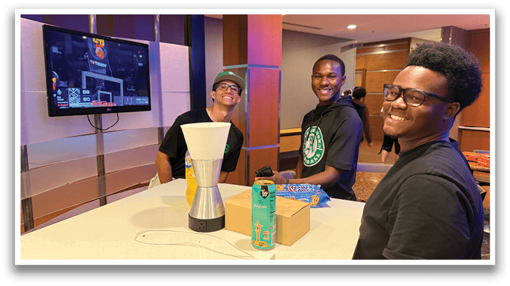 Three men are sitting at a counter, smiling and enjoying their time together. They are surrounded by various items, including a TV, a coffee maker, and a box of donuts. The men are wearing black shirts and glasses, and they appear to be having a good time in a casual setting. AI generated content