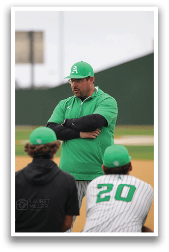 A man wearing a green shirt and a hat stands on a baseball field, looking at his players. AI generated content