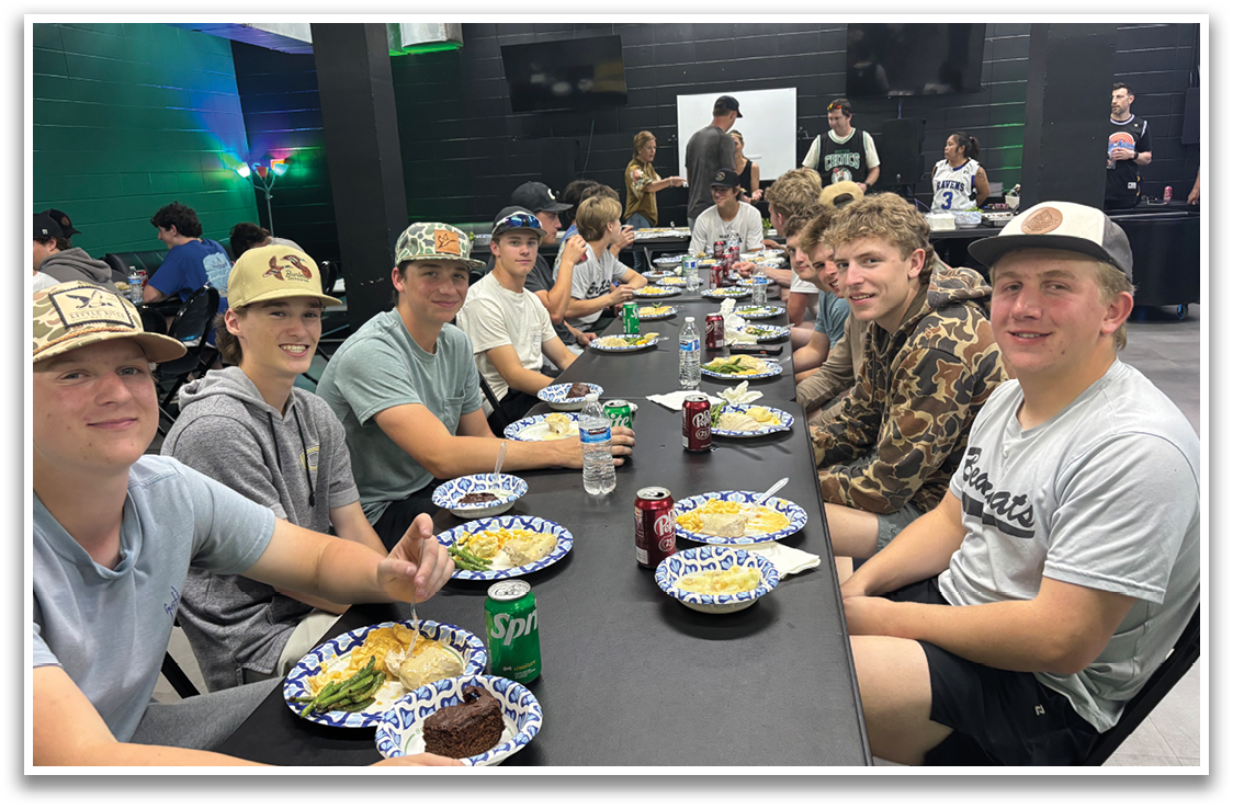 A group of young men are sitting around a table, eating and drinking. They are wearing baseball caps and are enjoying their meal together. AI generated content