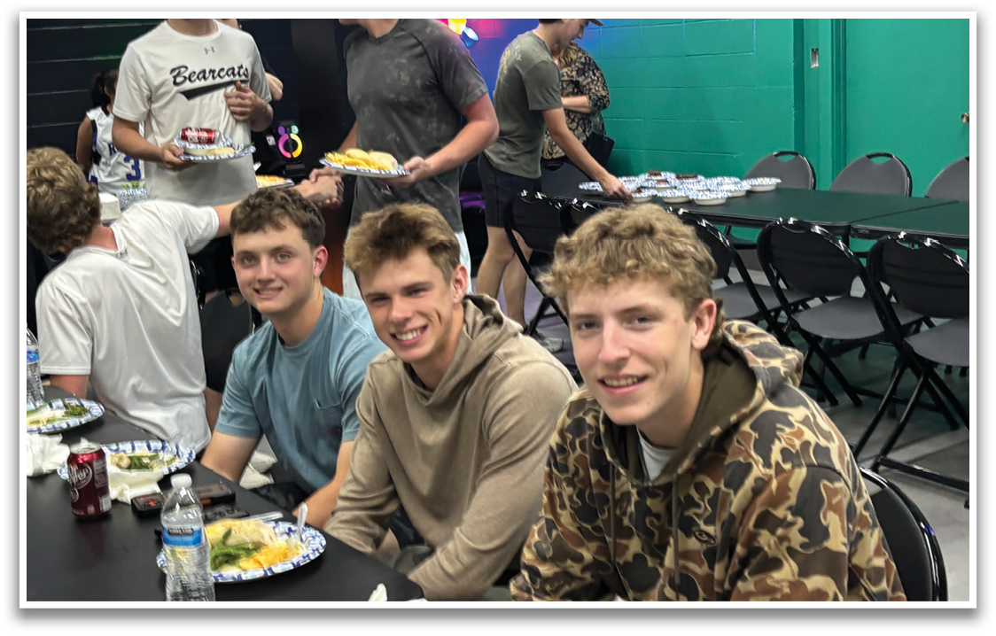 Three young men are sitting at a table with plates of food in front of them. They are smiling and posing for a picture. AI generated content