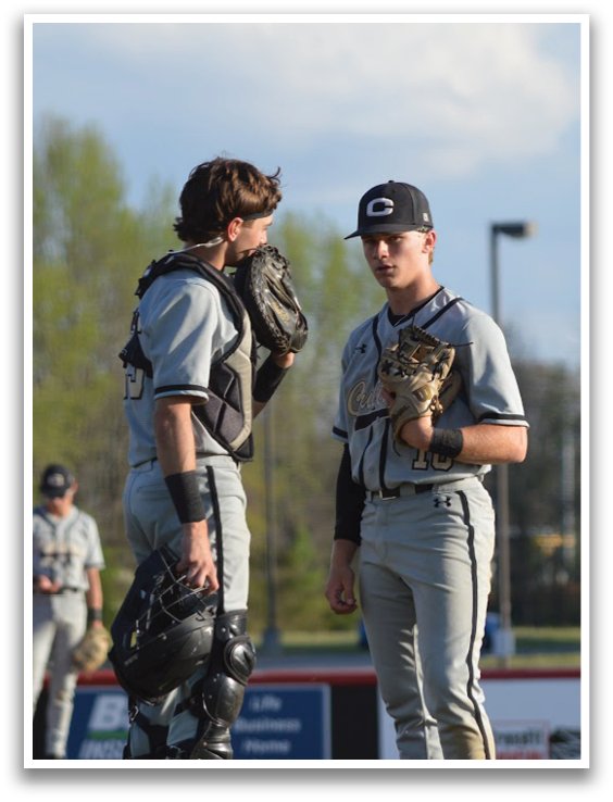 Two baseball players wearing grey uniforms and black gloves are standing on a field. One player is holding a baseball glove, while the other is holding a baseball. They are engaged in conversation, possibly discussing the game or their strategies. AI generated content