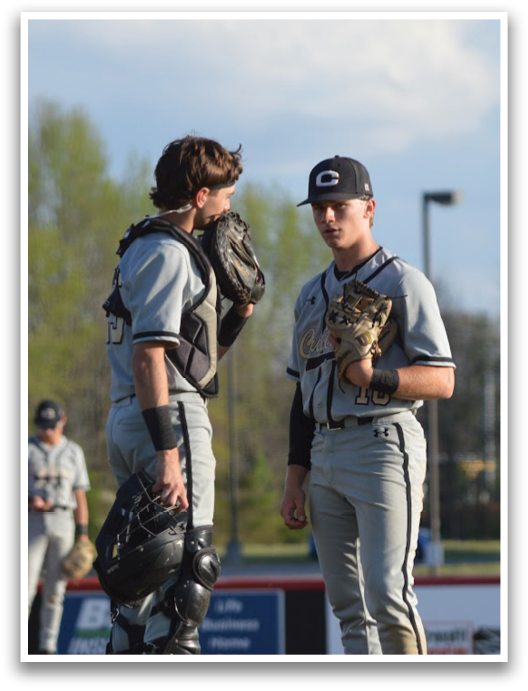 Two baseball players wearing grey uniforms and black gloves are standing on a field. One player is holding a baseball glove, while the other is holding a baseball. They are engaged in conversation, possibly discussing the game or their strategies. AI generated content
