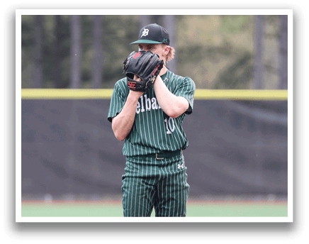 A baseball player in a green shirt and black pants is on the pitcher's mound. Description generated by AI