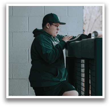 A man wearing a green hoodie and black shorts is leaning on a fence. Description generated by AI