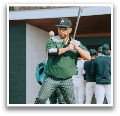 A man in a green jacket and white pants is holding a baseball bat and preparing to swing at a ball. Description generated by AI