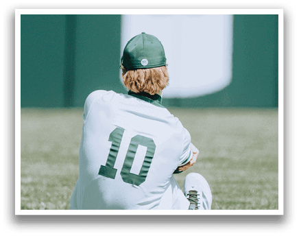 A baseball player wearing a green and white uniform sits on the grass. Description generated by AI