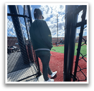 A man wearing a green jacket and white shoes is standing on a baseball field. Description generated by AI