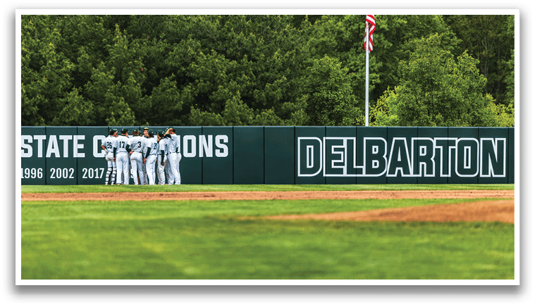 A group of baseball players standing in front of a green wall with the word Delaware on it. Description generated by AI