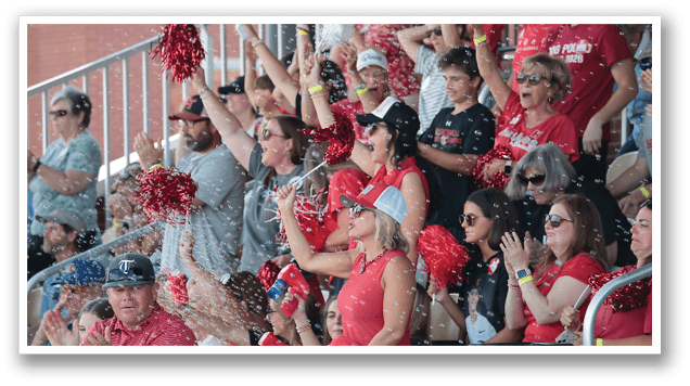 A group of people are sitting in the stands, watching a game. They are holding up red and white pom poms, cheering for their team. AI generated content