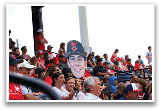 A crowd of people are sitting in the stands, watching a baseball game. A man is holding up a cardboard cutout of a baseball player, smiling. AI generated content