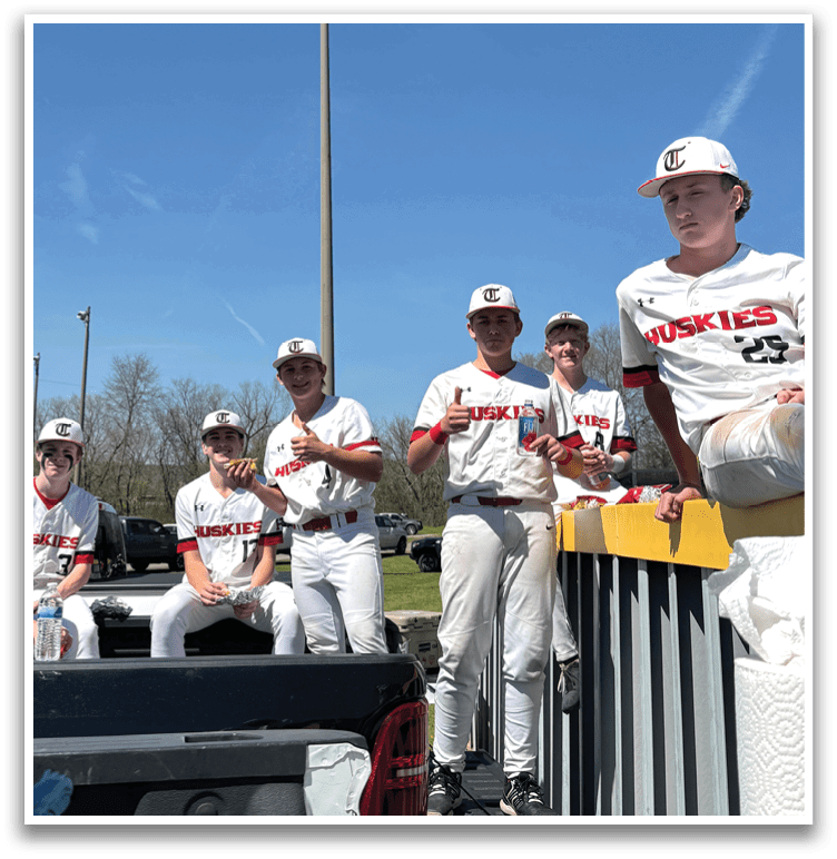 A group of baseball players are sitting on a truck, with some standing around it. They are wearing baseball uniforms and are likely taking a break from a game. AI generated content
