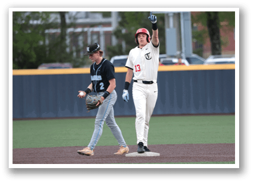 A baseball player wearing a red helmet and a white uniform is standing on a base, holding a bat. Another player is walking by, also holding a bat. There are several cars parked in the background. AI generated content