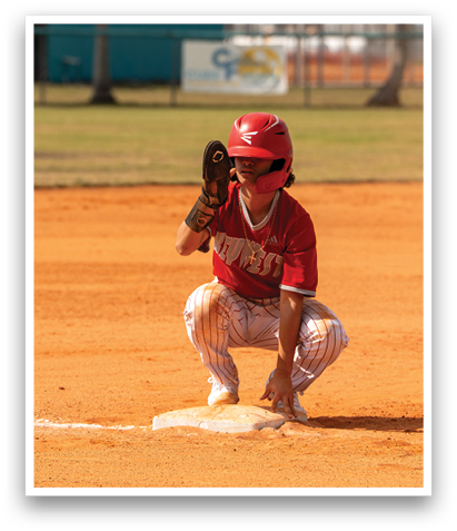 A young boy is kneeling on a baseball field, wearing a red shirt and a red helmet. He is holding a baseball glove, ready to catch the ball. The scene is set in a park with palm trees in the background. AI generated content