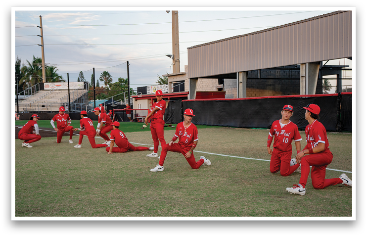 A group of young men wearing red shirts and white shoes are standing on a baseball field. They are all facing the same direction, possibly listening to a coach or waiting for a play to begin. AI generated content