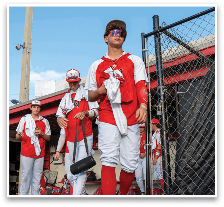 A group of baseball players are standing on the field, wearing red and white uniforms. One of the players is holding a baseball bat, and another player is holding a baseball glove. They are all standing in front of a fence, possibly waiting for their turn to play or practice. AI generated content