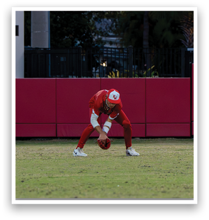 A baseball player in a red uniform is bending over to pick up a ball on a field. AI generated content
