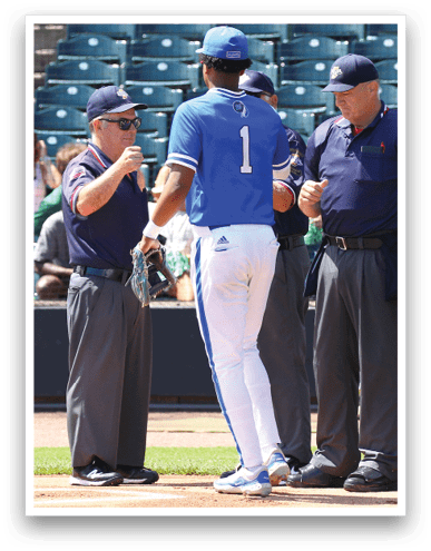 A person in a blue and white baseball uniform stands near three people in dark umpire uniforms on a baseball field. Description generated by AI