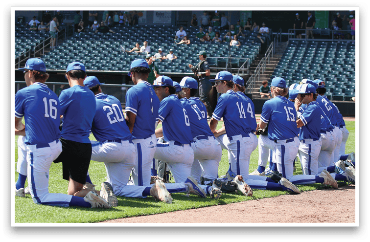 A group of people wearing blue baseball uniforms are kneeling in a row on the edge of a baseball field near the dugout. Description generated by AI
