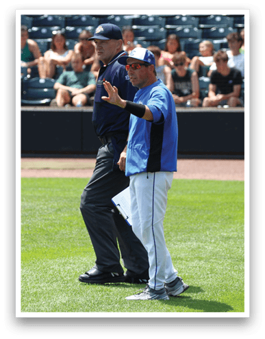 Two people in sports attire stand on a baseball field with one person gesturing with an open hand while spectators sit in the background. Description generated by AI