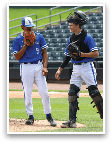 Two people in blue baseball uniforms are standing on a baseball field, with one wearing catcher's gear and holding a glove while the other holds a baseball glove near their face. Description generated by AI