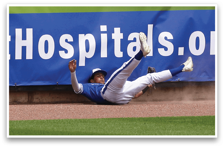 A person in a blue and white baseball uniform is falling backward on the ground near an outfield wall with a blue advertisement banner. Description generated by AI