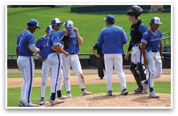 A group of people in blue baseball uniforms and one in catcher's gear are gathered on a pitcher's mound on a baseball field. Description generated by AI