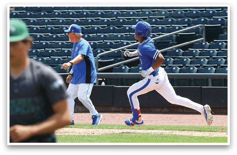 A person in a blue baseball uniform and helmet is running on a baseball field while another person in a blue shirt and cap walks nearby with empty stadium seats in the background. Description generated by AI