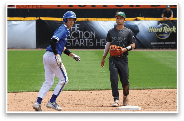 Two people in baseball uniforms are on a baseball field near a base, with one person wearing a blue uniform and helmet and the other wearing a dark uniform and green cap holding a baseball glove. Description generated by AI