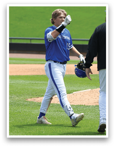 A person in a blue and white baseball uniform is walking on a grassy field holding a helmet and raising a gloved hand. Description generated by AI