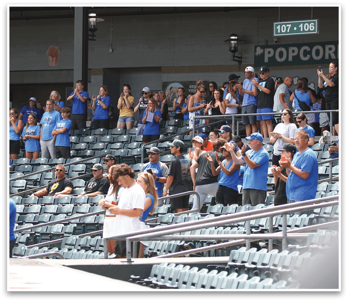 People in casual clothing are standing and clapping in stadium seating, with some seated and others using phones. Description generated by AI