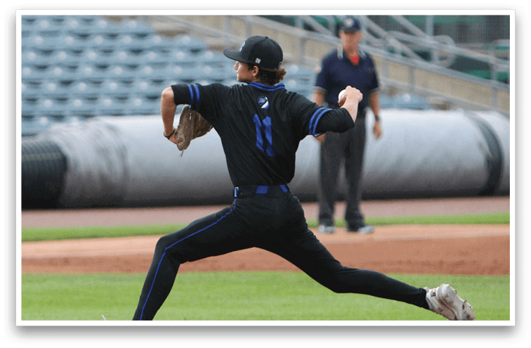 A person in a black sports uniform is pitching a baseball on a mound in an empty stadium with an official standing in the background. Description generated by AI