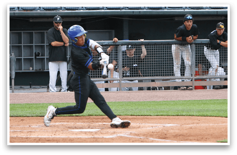 A person wearing a blue helmet and black sports uniform is swinging a baseball bat at a ball on a baseball field while several people in sports uniforms watch from behind a fence. Description generated by AI