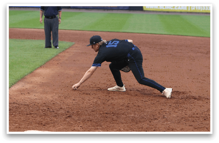 A person in a dark uniform is crouching and reaching to catch a baseball near a base on a baseball field while an official stands in the background. Description generated by AI