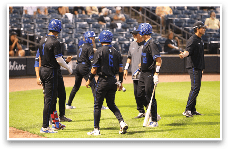 People wearing blue helmets and black sports uniforms are gathered on a baseball field near the dugout, with one person holding a bat and another person gesturing. Description generated by AI