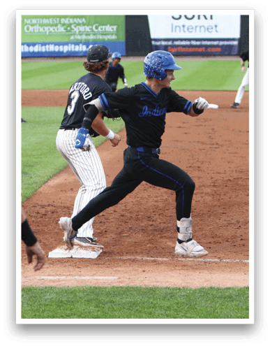 A person in a black and blue baseball uniform is running toward a base while another person in a black and white striped uniform stands nearby and two other players are positioned on the field. Description generated by AI