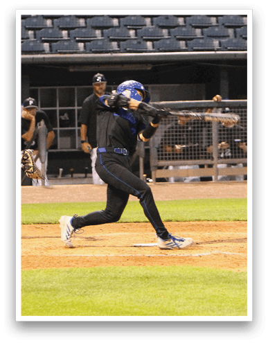 A person wearing a blue helmet and black sports uniform is swinging a baseball bat at a ball on a baseball field with other people in the background near a dugout. Description generated by AI