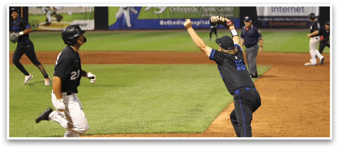 A person in a black and blue uniform is catching a baseball at first base while another person in a black jersey and white pants runs toward the base on a baseball field. Description generated by AI