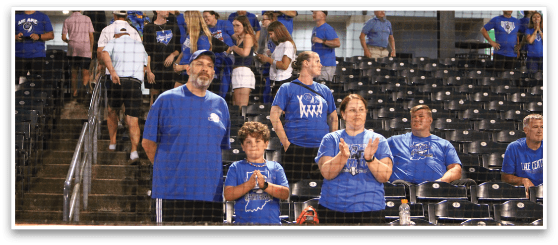 People wearing blue shirts are standing and clapping in a stadium with empty black seats and a “First Aid" sign visible in the background. Description generated by AI