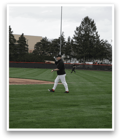 A man is playing baseball in a field, wearing a black shirt and white pants. He is holding a baseball bat and pointing it towards the ground. There are other people in the field, possibly teammates or opponents. The scene is set in a grassy area with a building in the background. AI generated content