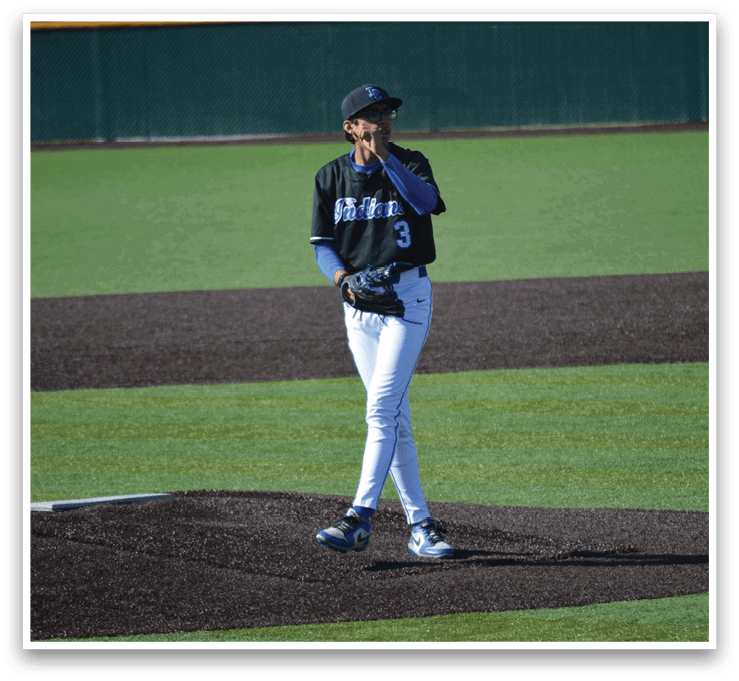A baseball player wearing a black shirt and white pants stands on the pitcher's mound. Description generated by AI