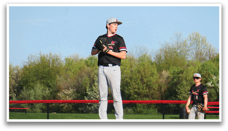 A baseball player in a black shirt and red shoes stands on a mound. Description generated by AI